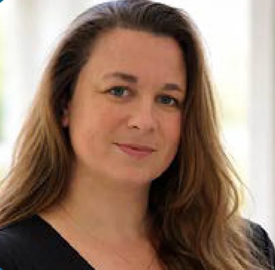 Portrait of a woman with long brown hair wearing a black top, smiling slightly at the camera against a light background